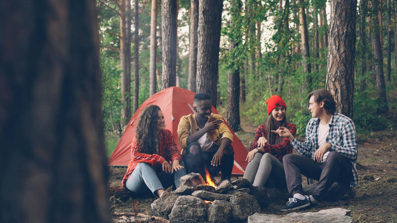 Four friends sitting around a campfire in a forest with a red tent in the background.