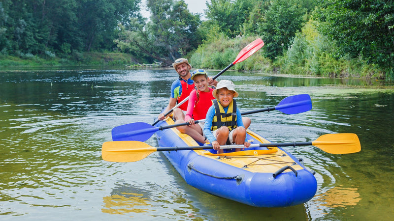 Three children paddling a blue kayak on a calm river with trees in the background